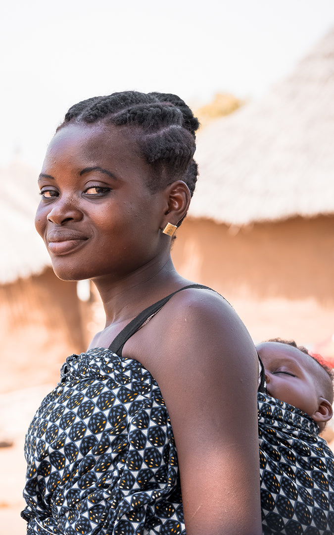 A Ghanaian woman carries her child on her back using a sling. The child is sleeping. She has her hair braided and tied in a bun.