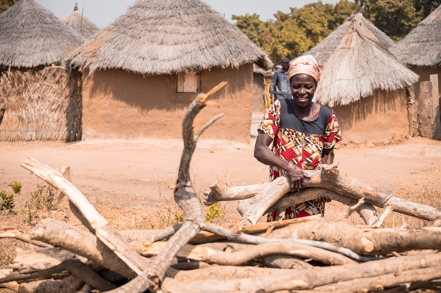 A Ghanaian woman is smiling as she collects firewood. Behind her are several Ghanaian houses. There is also a man in the background, wearing black clothes and a black cap.