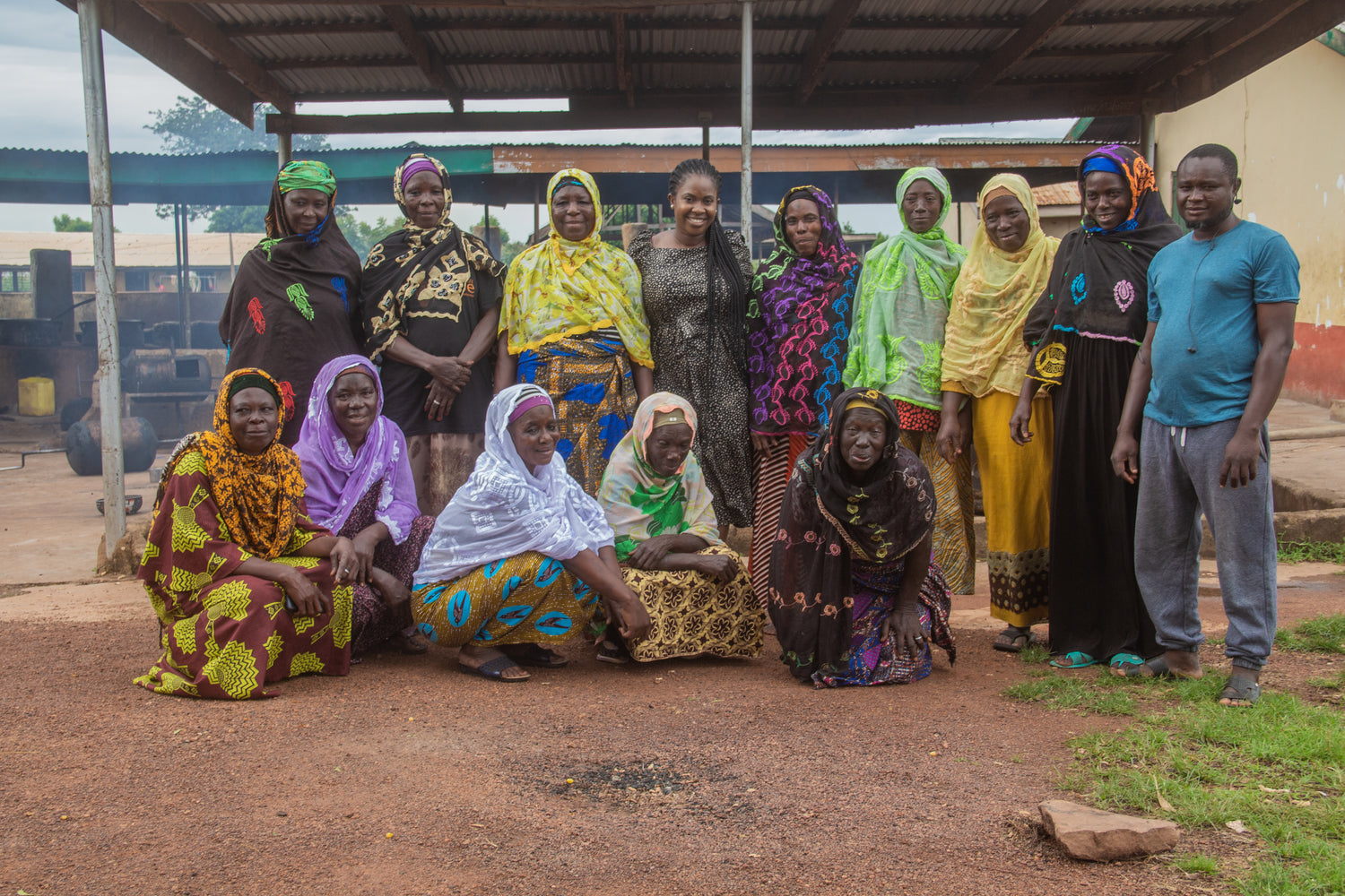 A group of Ghanaian women are in position for a group photo, some are standing in the back row and others are seated in the front row. They wear traditional Ghanaian clothing and head scarves. Among them is Mildred the founder of Mimaami Orgnaics.