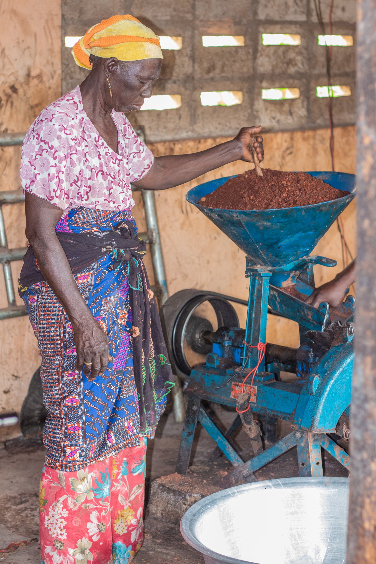 A Ghanaian woman is processing Shea on a machine. She is standing wearing traditional clothes and a headscarf. With her left hand she uses a pestle to push the Shea into the processor.