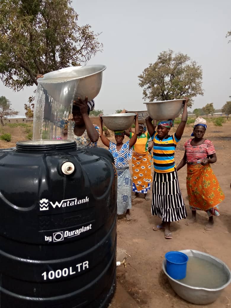 A group of five Ghanaian women are carrying water into containers. Some of them carry water in basins over their heads. All wear traditional regional clothes and turbans.