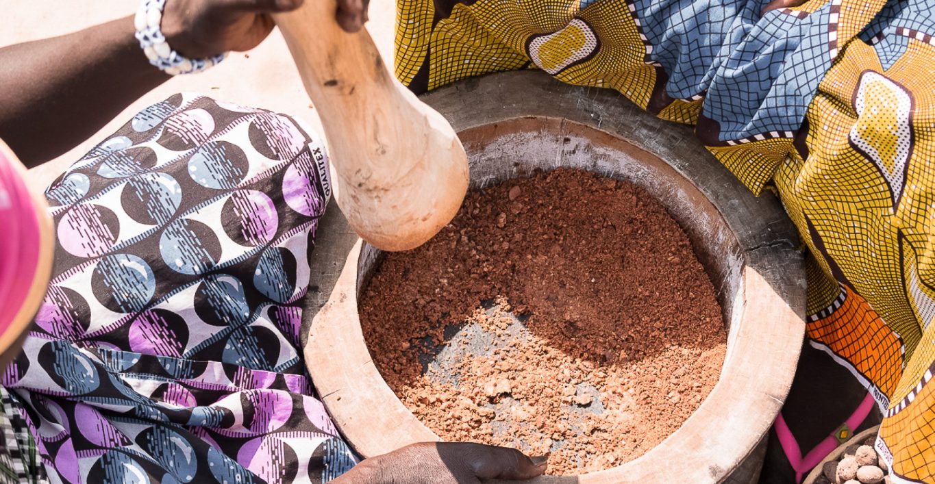 A Ghanaian woman is grinding a shea nut using a pestle. The pestle is resting on her legs.